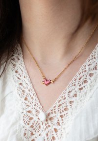 Zlata verižica z obeskom v obliki roza srca, prebodenega z puščico, nošena čez belo čipkasto bluzo s tkaninskimi gumbi.