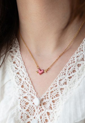 Złoty łańcuszek z różowym sercem przebitym strzałą, noszony na białej koronkowej bluzce z guzikami obłożonymi tkaniną.