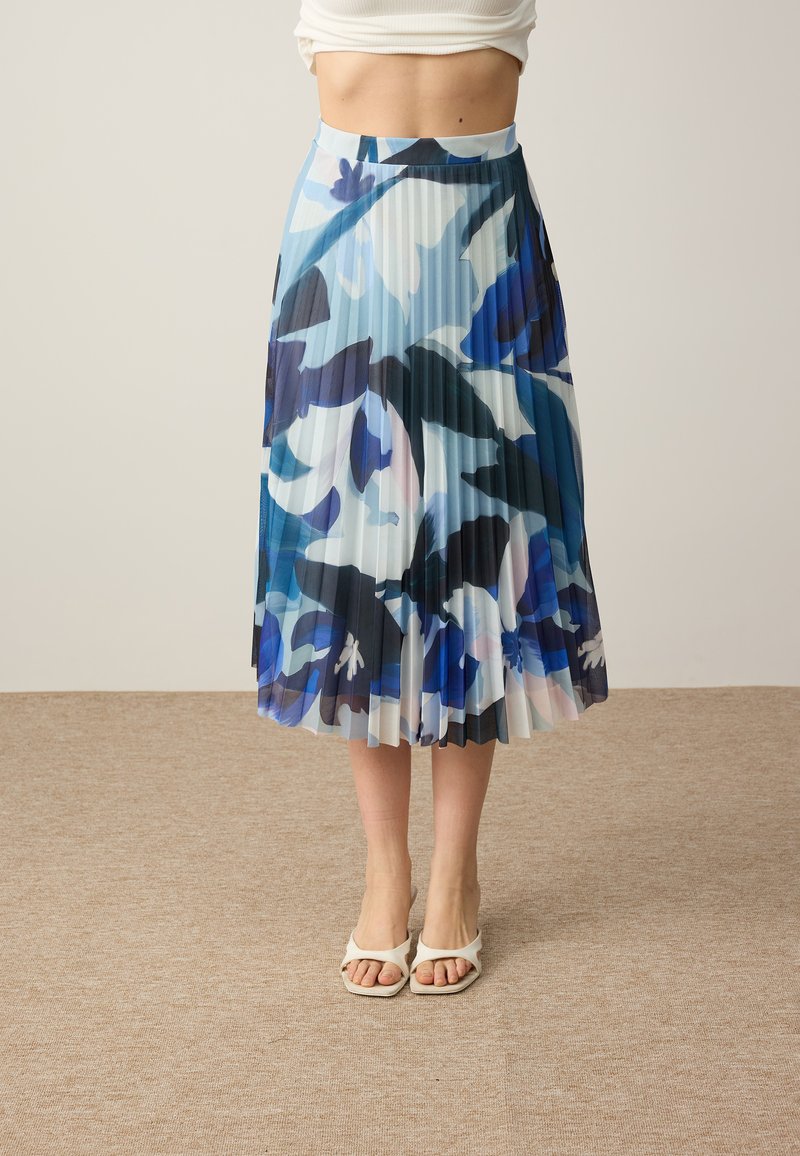 Femme portant une jupe midi plissée taille haute avec des motifs floraux bleus, noirs et blancs et des sandales blanches à bout ouvert.