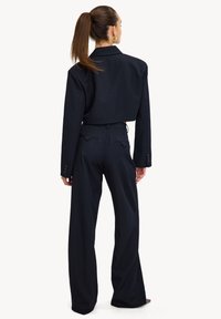 Blazer cropped bleu marine aux épaules structurées, associé à un pantalon large taille haute. Tissu lisse avec des détails subtils sur les poches arrière.