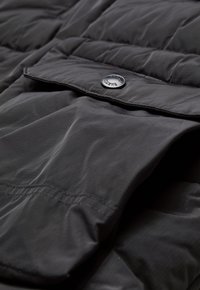 Black padded jacket detail featuring a secure flap pocket and a branded snap button, showcasing smooth, durable fabric texture and stitching.