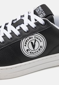 Černé kožené tenisky s bílými tkaničkami a texturovaným povrchem. Obsahují kruhové logo s textem "VERSACE JEANS COUTURE" v černé a bílé barvě. Bílá podrážka.