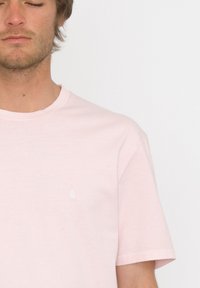 Vaaleanpunainen puuvillainen t-paita, jossa on lyhyet hihat, pyöreä kaula-aukko ja pieni valkoinen logo vasemmassa rinnassa. Sileä pinta, rento malli.