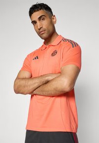 Polo shirt corail avec col, présentant des accents rayés gris foncé sur les épaules et un détail de logo. Le tissu semble texturé et respirant.