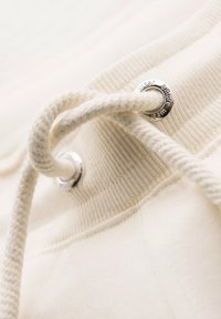 Détail d'un hoodie de couleur crème montrant un cordon blanc traversé par un œillet en métal argenté, avec une texture côtelée autour de la base.
