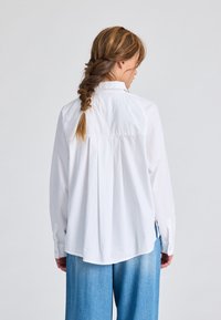 Camicia bianca a maniche lunghe con una vestibilità rilassata, caratterizzata da una piega sul retro, un orlo arrotondato e polsini con bottoni, abbinata a pantaloni in denim azzurri.