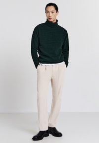 Dunkelgrüner Rollkragenpullover, hellbeige strukturierte Hose, schwarze Knöchelstiefel. Schlichte Linien, lockere Passform, einfarbige Farben, minimale Details.