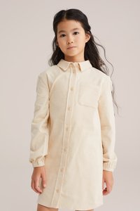 Ilgi piedurkņu, ar apkakli tērps gaiši beidžā kordūrā ar priekšējām pogām un krūšu kabatu; ietver savilktus cuffs papildu detaļu pievienošanai.