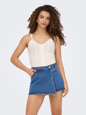 Jeune femme aux cheveux bouclés portant un débardeur en maille blanche et une jupe en jean bleue avec des motifs de cœurs rouges, posant avec une main dans la poche.