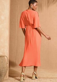 Koraal mididress met brede mouwen, gathered taille en een gladde textuur. Gecombineerd met goudkleurige sandalen met blokhak en bandjes.
