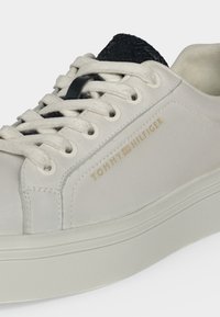 Gros plan d’une basket blanche avec une semelle épaisse, des lacets blancs, une languette noire texturée et un logo Tommy Hilfiger doré sur le panneau latéral.