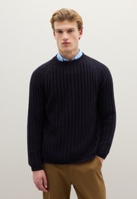 Marineblauer Strickpullover mit Rippenstruktur, rundem Ausschnitt und langen Ärmeln. Kombiniert mit einem hellblauen Hemd mit Kragen darunter.