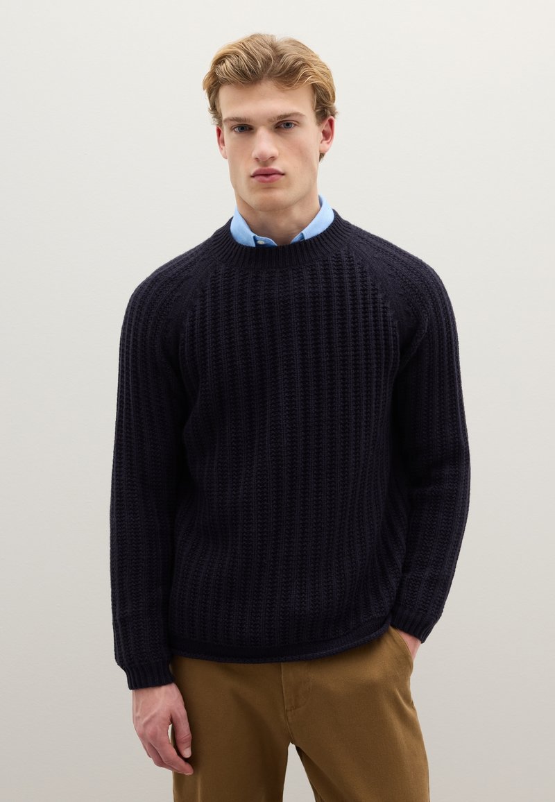 Marineblauer Strickpullover mit Rippenstruktur, rundem Ausschnitt und langen Ärmeln. Kombiniert mit einem hellblauen Hemd mit Kragen darunter.