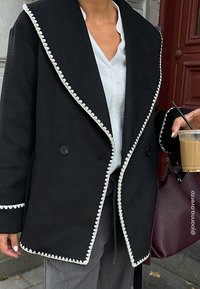 Czarny wełniany płaszcz z dużym kołnierzem szalowym, białym falbanym wykończeniem, podwójnymi guzikami i kieszeniami, noszony na białej koszuli i szarych spodniach.