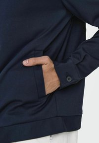 Marineblå sweatshirt laget av mykt stoff, med sidelomme og knappedetalj på mansjettene, med en avslappet passform og jevn tekstur.