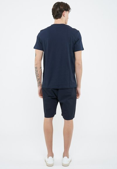 Uomo con capelli castano corti, t-shirt blu navy e pantaloncini, in piedi rivolto di spalle, che mostra un tatuaggio floreale sul braccio sinistro, indossa scarpe casual bianche.