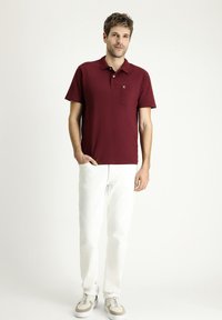 Polo shirt bordeaux a maniche corte con tasca frontale e branding sottile, abbinato a jeans bianchi a gamba dritta e scarpe da ginnastica con dettagli color beige.