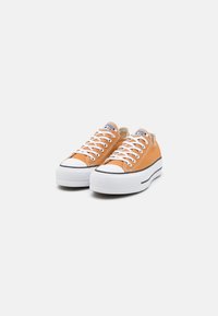 Ett par låga orange canvas-sneakers med vita gummisulor och vita snören, uppställda på en enkel vit bakgrund.