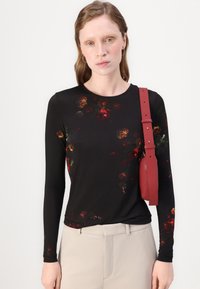 Langärmliges schwarzes Top mit einem floralen Muster in Rot und Grün, kombiniert mit beigen Hosen und einer roten Schultertasche mit einem Tragegurt.