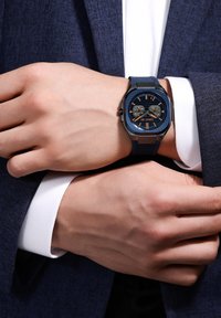 Zils analogais pulkstenis ar teksturētu gumijas siksnu, astoņstūra korpusu un kontrastējošiem oranžiem akcentiem uz tumša ciparnīcas. Valkāts uz rokas.