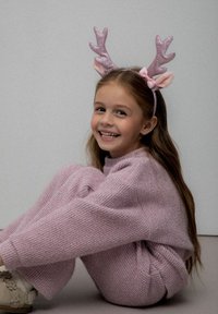 Różowy, dziany sweter i spodnie, miękka tekstura, noszone z różową opaską na głowie z błyszczącymi rogami i uszami. Dziecko siedzi, uśmiechając się.