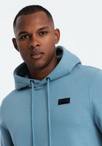 Sudadera azul claro con superficie texturizada, que presenta un bolsillo tipo canguro y un parche rectangular negro con logotipo en el pecho. Los cordones tienen puntas negras.