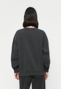 Persona con capelli scuri fino alle spalle, che indossa una felpa grigia antracite oversize e pantaloni coordinati, in piedi di fronte a un muro bianco.