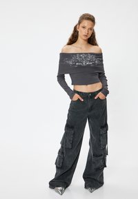 Graues, schulterfreies, geripptes Oberteil mit grafischem Design, kombiniert mit lockeren, dunkelgrauen Cargo-Hosen mit mehreren Taschen und silbernen, spitzen Schuhen.