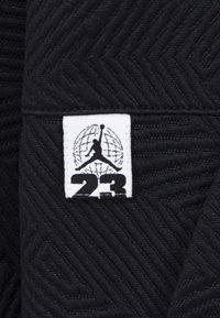 Tessuto nero intrecciato con una texture a zigzag. Presenta un'etichetta bianca con il logo Jumpman e "23" sotto, centrata.