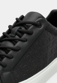Primo piano di una sneaker in pelle nera con iniziali del marchio impresse di lato e spesse stringhe nere su una suola bianca.