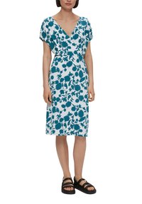 Robe à imprimé floral en teal sur fond blanc. Encolure en V, manches dolman courtes, coupe jusqu'au genou, associée à des sandales noires à lanières.