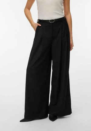 Frau trägt hoch-taillierte, schwarze, weit geschnittene Hosen mit Gürtel, kombiniert mit einem weißen ärmellosen Top und schwarzen spitzen Schuhen.