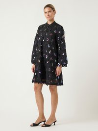 Γυναίκα που φοράει ένα μαύρο φόρεμα έως το γόνατο με ιριδίζοντες κυκλικούς παγιέτες και μακριά μανίκια, συνδυασμένο με μαύρες ανοιχτές, ψηλοτάκουνες γόβες.