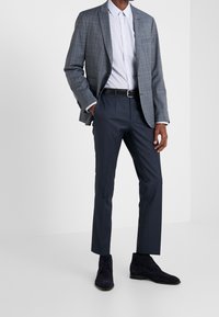 Grau kariertes Blazer über einem hellen gemusterten Hemd, kombiniert mit dunklen Hosen und schwarzen Wildleder-Ankle-Boots. Schmaler Schnitt und maßgeschneiderte Gestaltung.