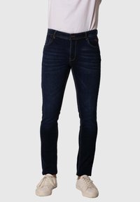 Vaqueros de denim de lavado oscuro con un corte ajustado, que presentan costuras en contraste, cinco bolsillos y un cierre de botón, combinados con zapatillas blancas.