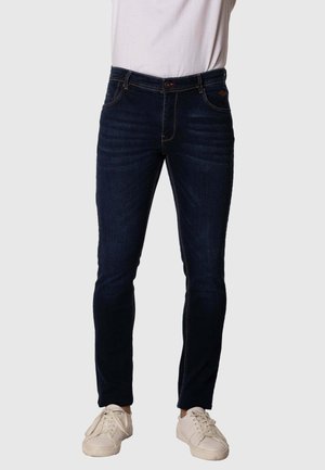 Vaqueros de denim de lavado oscuro con un corte ajustado, que presentan costuras en contraste, cinco bolsillos y un cierre de botón, combinados con zapatillas blancas.