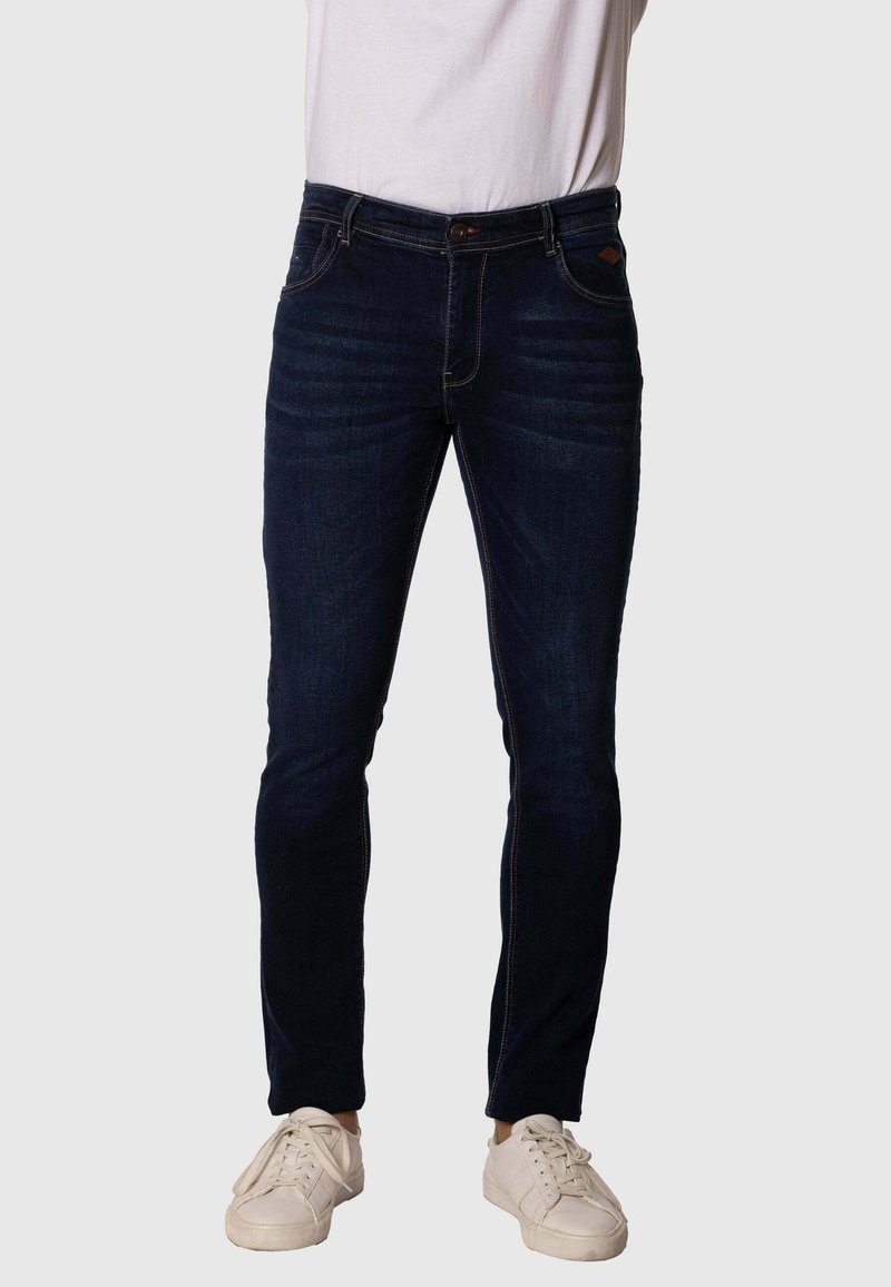 Vaqueros de denim de lavado oscuro con un corte ajustado, que presentan costuras en contraste, cinco bolsillos y un cierre de botón, combinados con zapatillas blancas.