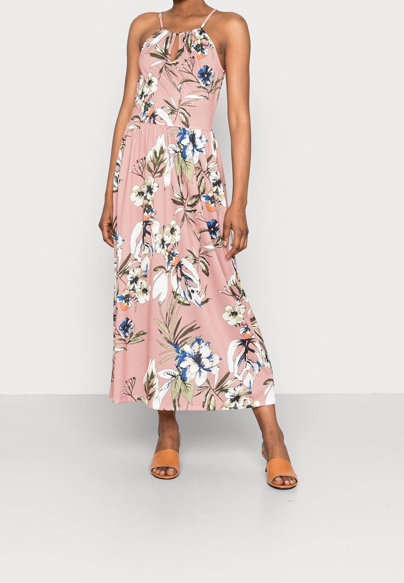 Femme portant une robe longue à bretelles rose à fleurs avec un décolleté en forme de trou de serrure et des talons ouverts couleur beige, debout devant un fond uni.