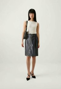 Mulher com cabelo preto longo, vestindo uma blusa branca sem mangas, saia lápis de couro preta com zíper frontal, bolsa preta e saltos pretos.