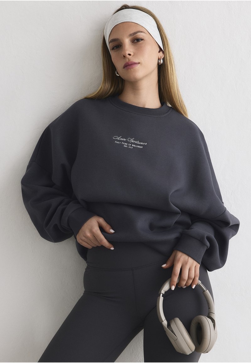 Jeune femme en tenue de sport sombre et bandeau, appuyée contre un mur, tenant un casque beige couvrant les oreilles dans une main.