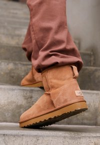 Botas de tobillo de ante beige con punta redondeada, detalles de costura y suela de goma, combinadas con pantalones rosas de corte holgado en escaleras de concreto.