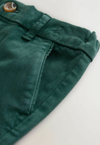 Tissu en velours côtelé vert avec une fermeture à bouton, présentant une taille pliée et une ouverture de poche sur le côté. Texture lisse et design épuré.
