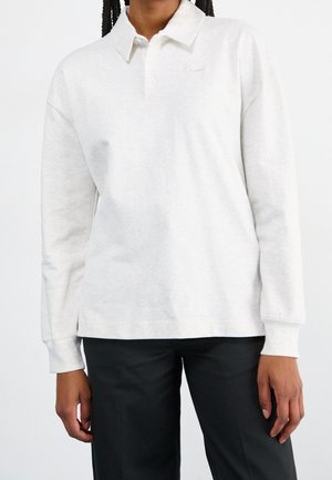 Osoba ubrana w białą, długorękawową koszulkę polo z kołnierzykiem i czarne spodnie, stojąca na jednolitym białym tle.