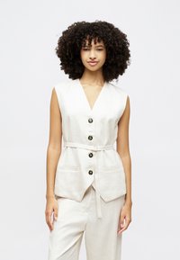 Gilet en lin beige avec un col en V, quatre boutons noirs et une taille à nouer. Il dispose de poches latérales et d'une silhouette ajustée avec un design sans manches.