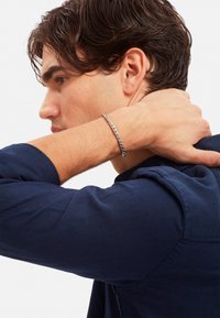 Bracciale in metallo argentato con design intrecciato, indossato su un polso, su una camicia blu navy. Presenta una texture liscia e una superficie riflettente.