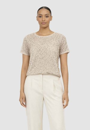 Donna che indossa una maglia beige a maniche corte con paillettes abbinata a pantaloni a vita alta color crema plissettati, in piedi davanti a uno sfondo semplice.