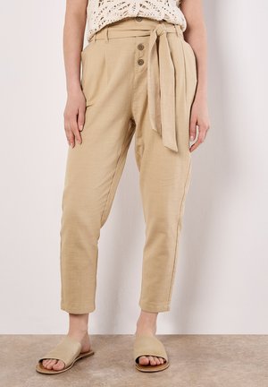 Modella che indossa pantaloni beige a vita alta con una cintura, abbinati a sandali a scivolo neutri, si trova davanti a uno sfondo semplice.