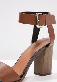 Sandale à talon en cuir marron avec une large bride et une boucle au niveau de la cheville. Dotée d'un talon bloc avec une apparence de bois texturé. Semelle intérieure lisse.