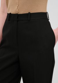 Černé tailored kalhoty s hladkou texturou, postranními kapsami a poutky na opasek, doplněné o žebrovaný béžový top. Knoflíky dodávají detail.