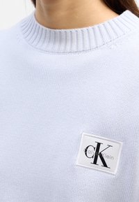 Personne portant un pull en tricot bleu clair avec un col rond côtelé et un petit patch rectangulaire avec le logo Calvin Klein sur la poitrine.